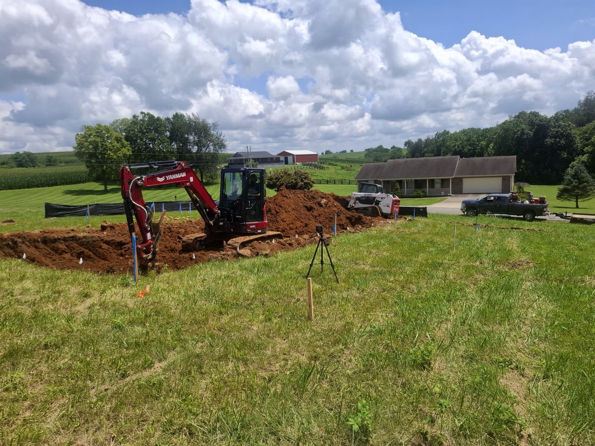 Excavator digging a foundation trench on a rural Kentucky building site with a laser level set up on a tripod and grade stakes in the ground